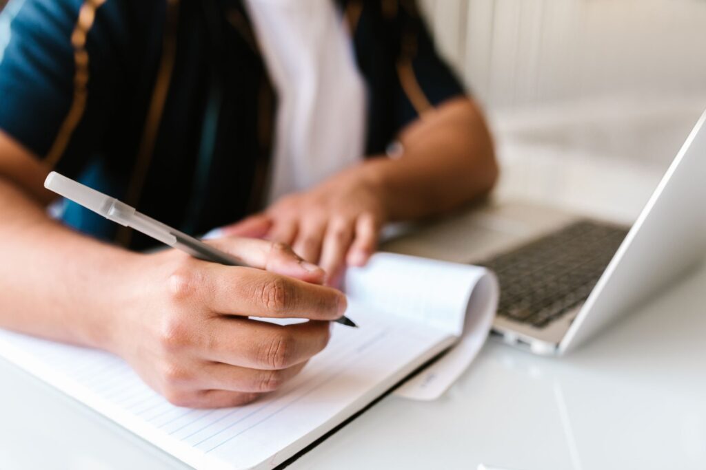 Focused image of a person writing in a notebook beside an open laptop